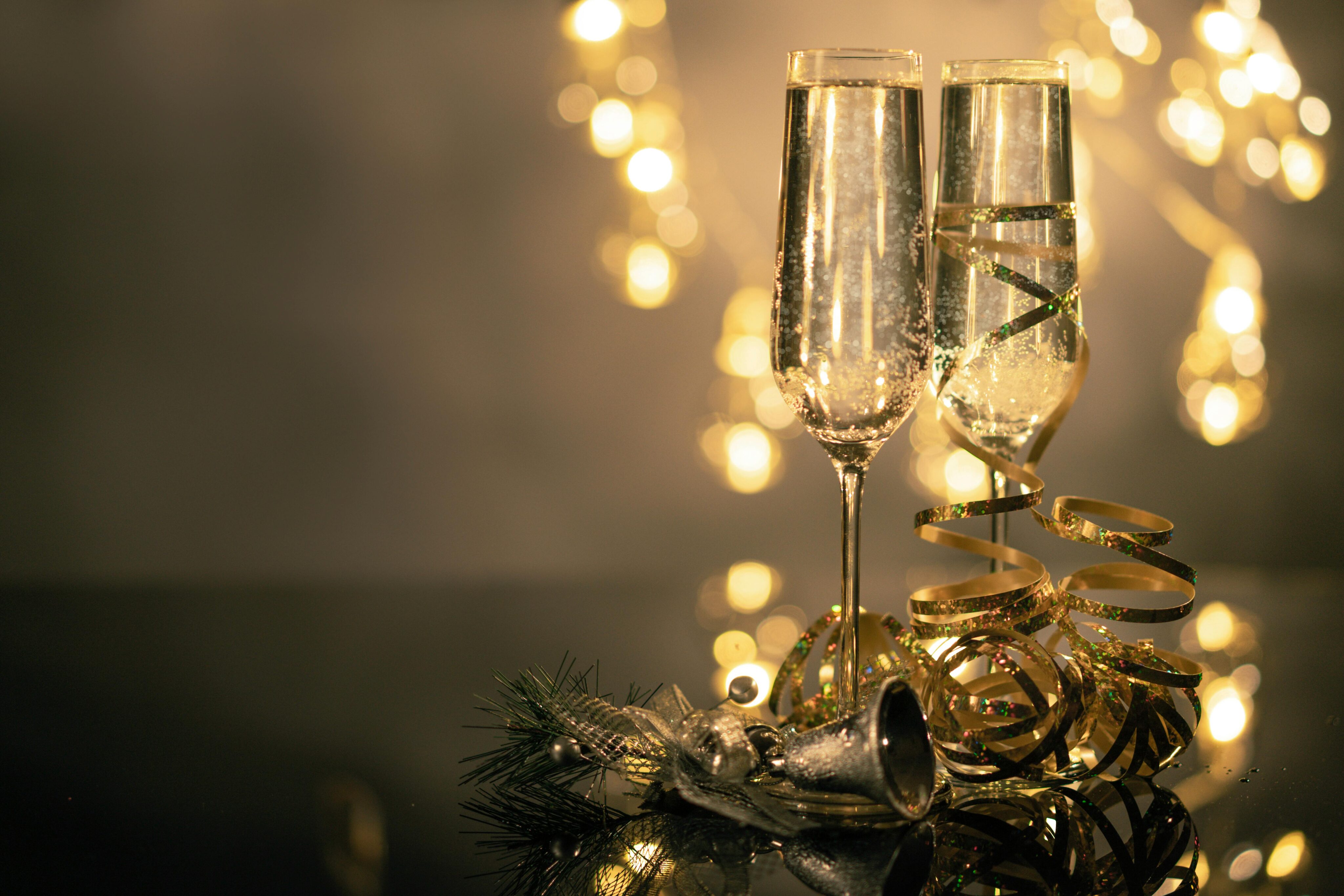 Two champagne flutes filled with bubbly liquid are adorned with golden ribbon on a reflective surface, capturing the essence of New Year's Eve. A small silver bell and pine branch are placed nearby, creating a festive ambiance with blurred lights in the background.