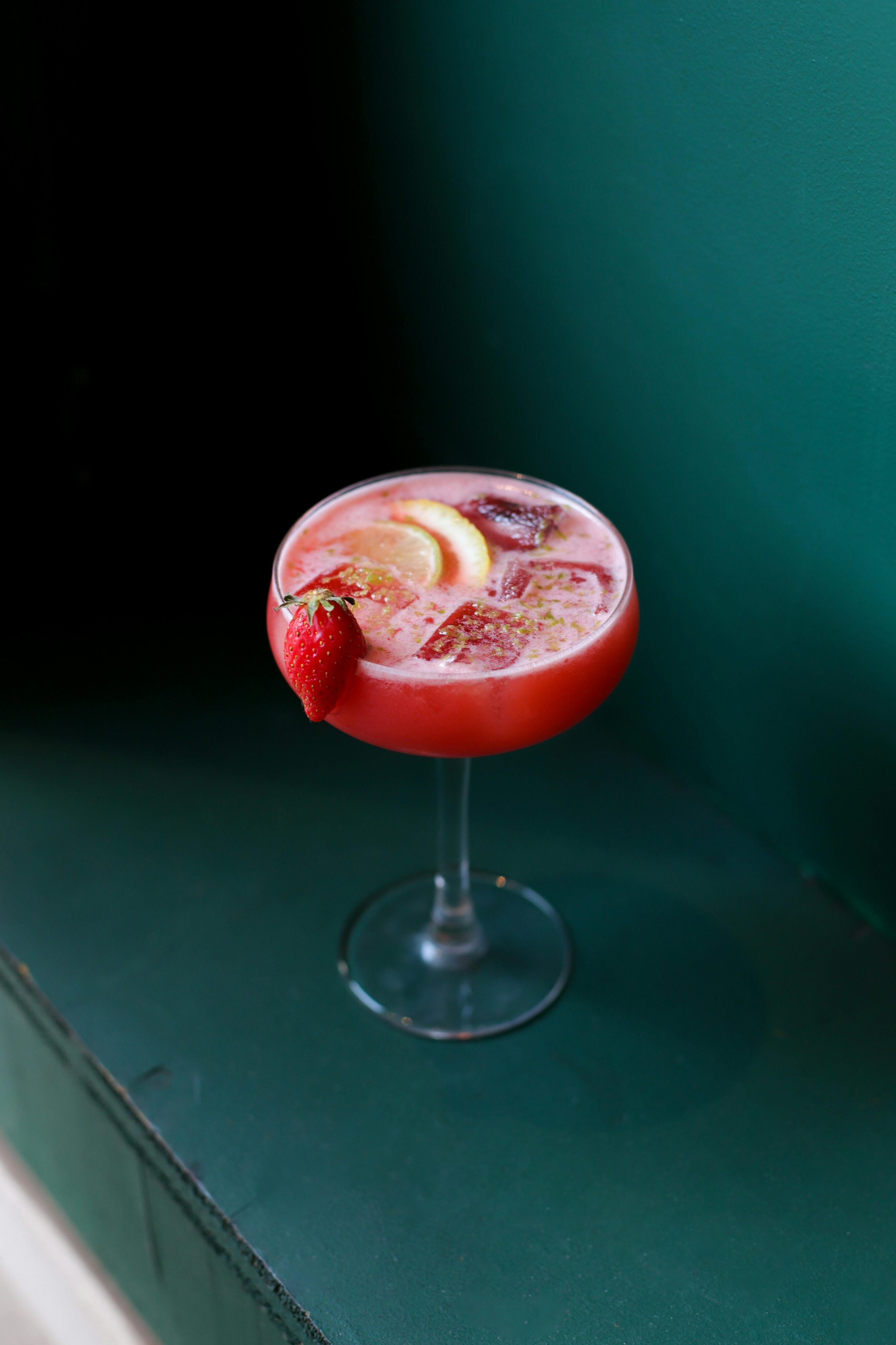 A pink cocktail in a glass garnished with a lemon slice, strawberry, and ice cubes, set against a dark green background—perfect for February cocktail specials.