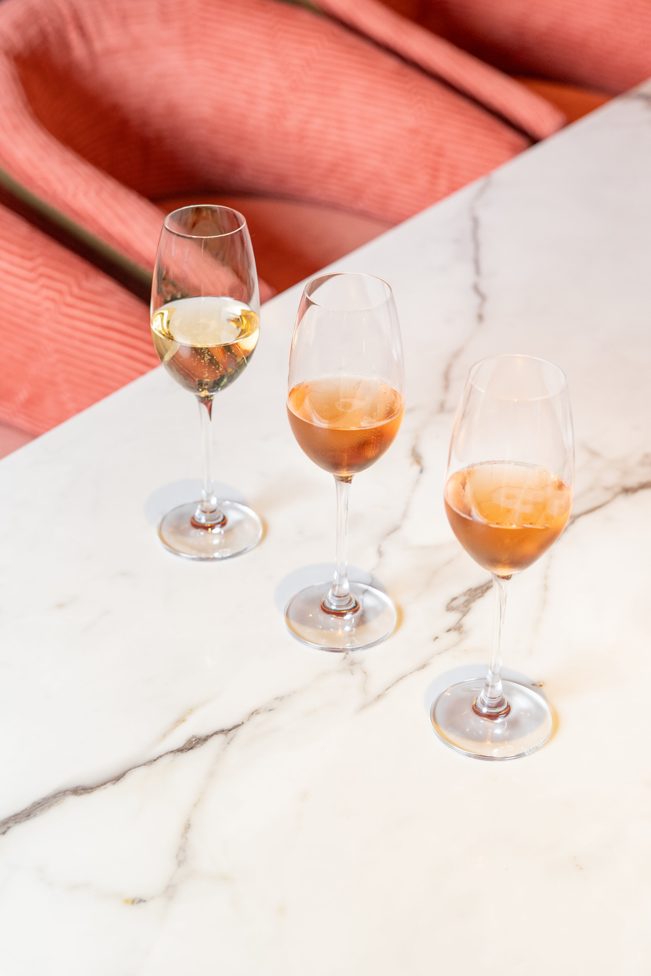 Three wine glasses with different types of wine are placed on a white marble table, with a pink textured fabric background.