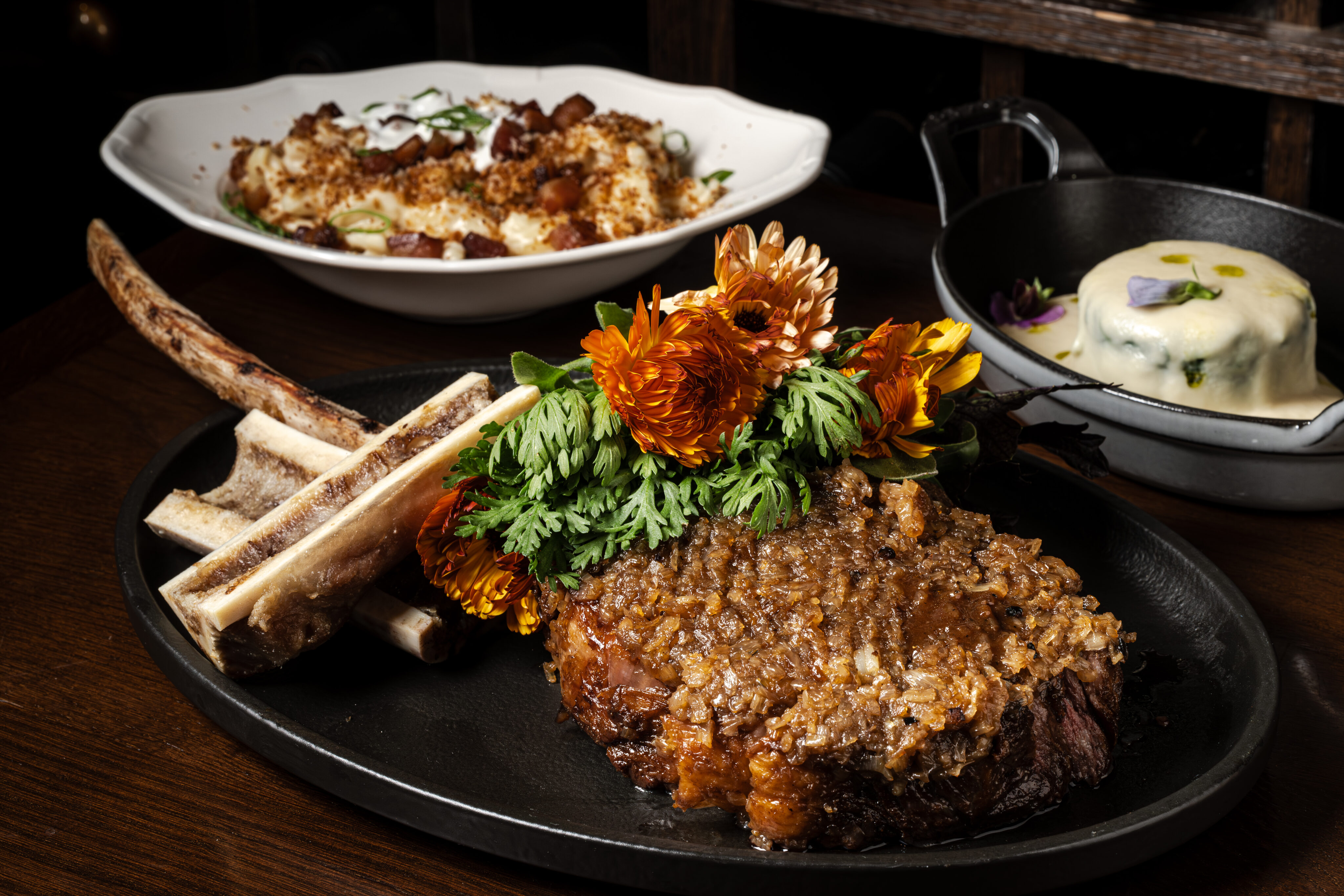A plated steak garnished with edible flowers and herbs, served with bone marrow, is the star of this dinner; sides of vegetables and a covered dish complete the menu in the background.