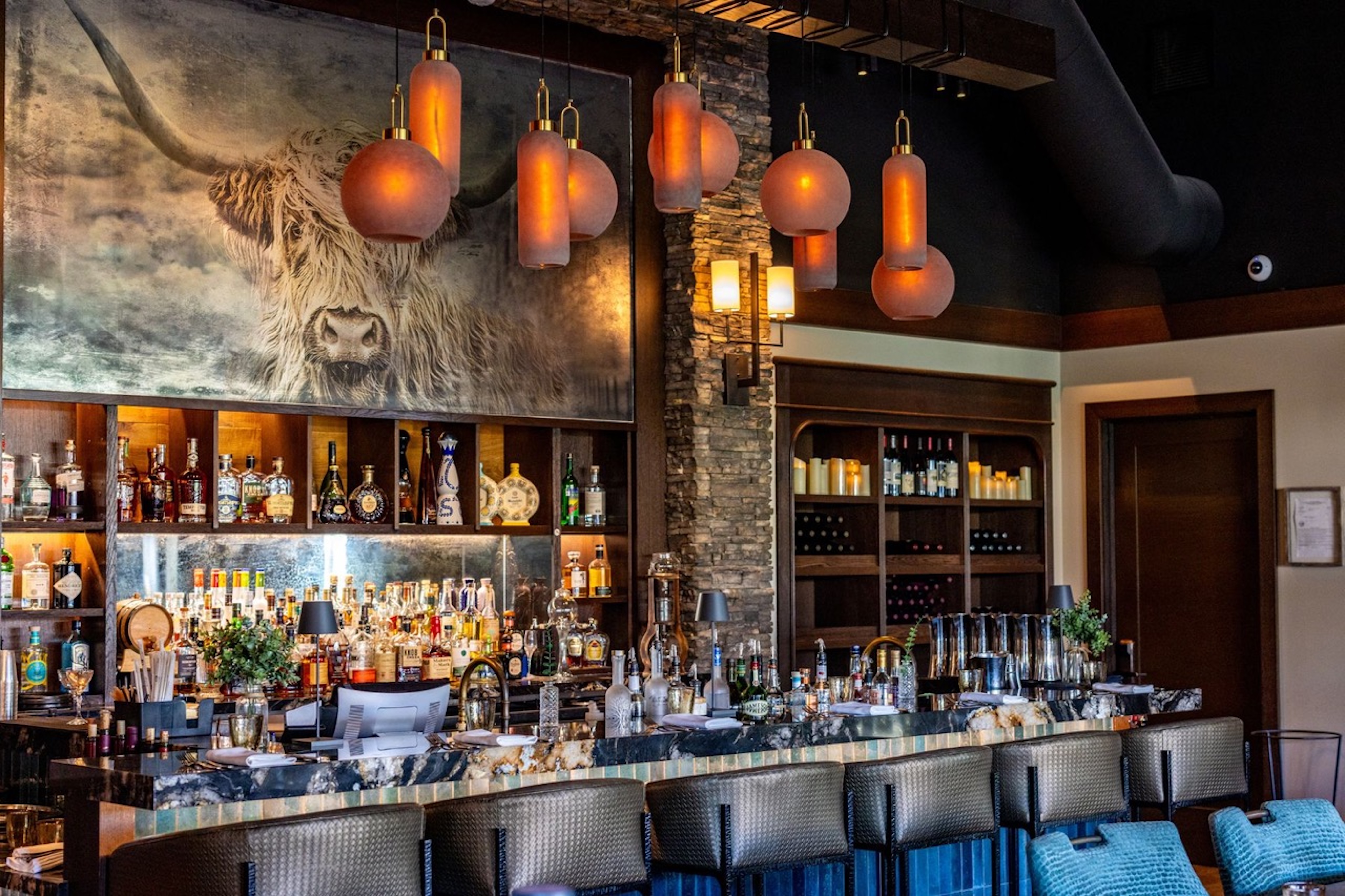 A modern bar with a stone accent wall, shelves of liquor and wine, a marble countertop, blue bar stools, and a large painting of a highland cow—part of the Wild Game Series—showcasing the spirit and hooves of the wild above the bar.