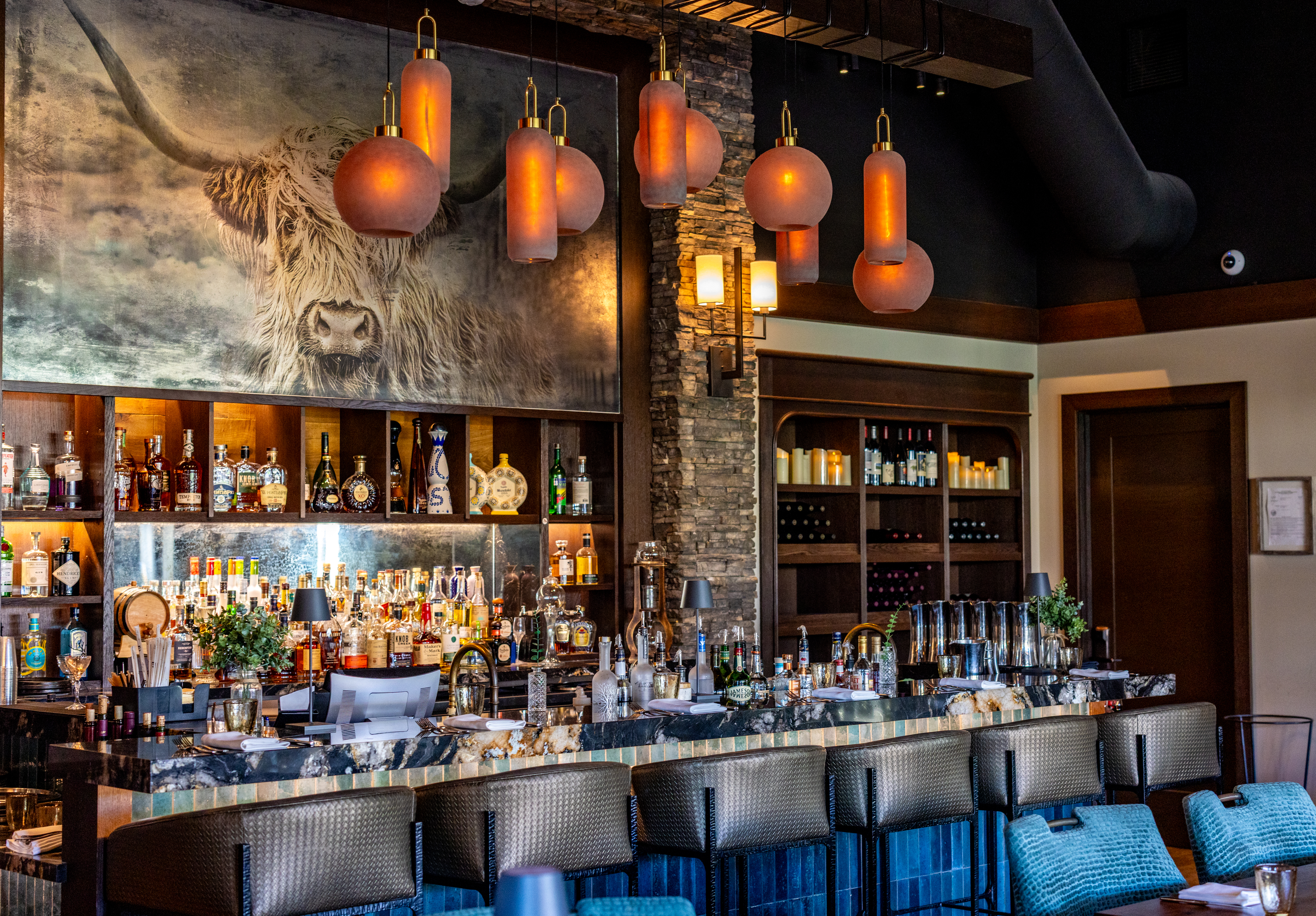 Modern bar interior with stone accents, shelves of liquor and wine, blue bar stools, and a large painting of a highland cow on the wall—perfect for a stylish New Year's Eve Dinner to welcome 2025. Warm pendant lights hang above the counter.