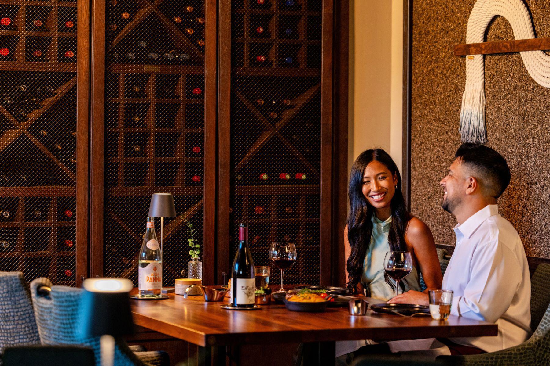 Two people sit at a restaurant table with wine and food, smiling and talking. Behind them is a large wine rack filled with bottles—perfect for celebrating a special Black Friday offer on fine wine.