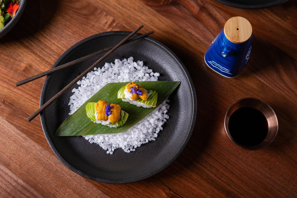 Two pieces of sushi from the Wild Game Series, with yellow topping and edible flowers on rice, are served on a leaf over coarse salt in a black dish, accompanied by chopsticks, soy sauce, and a blue sake bottle nearby.