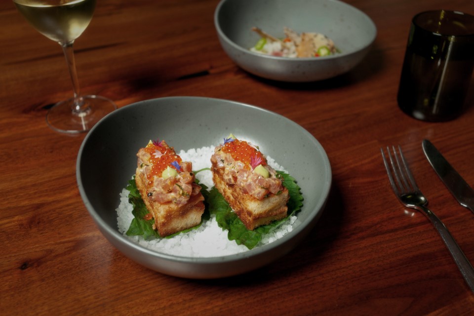 Two pieces of toast topped with diced fish and orange roe on a green leaf, served in a gray bowl with a white powder base; part of the Wild Game Series, with a wine glass and utensils nearby on a wooden table.