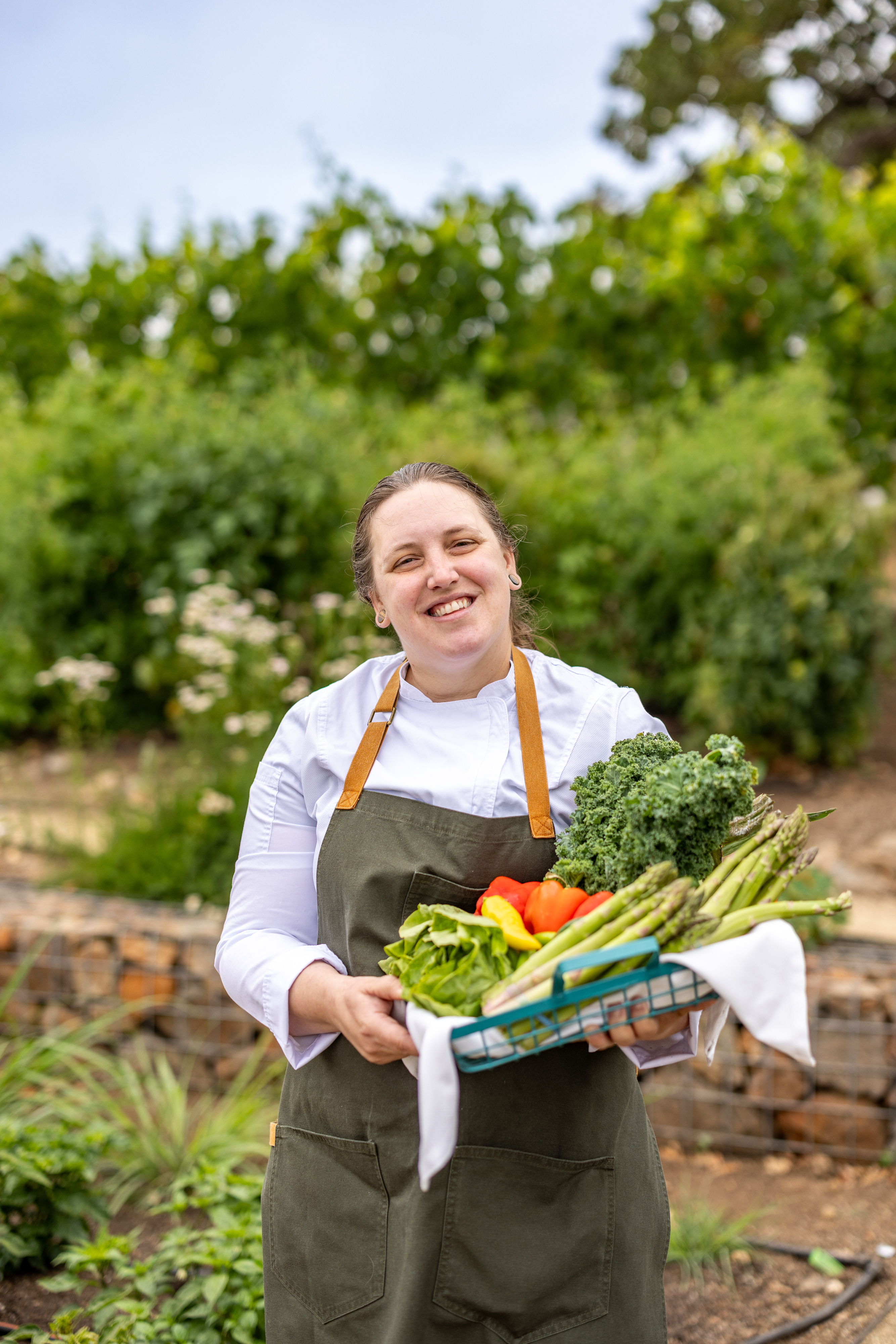 A woman in a chef's uniform and apron stands outdoors holding a basket of fresh vegetables, ready to prepare a dish for the Wild Game Series, featuring kale, bell peppers, and asparagus.