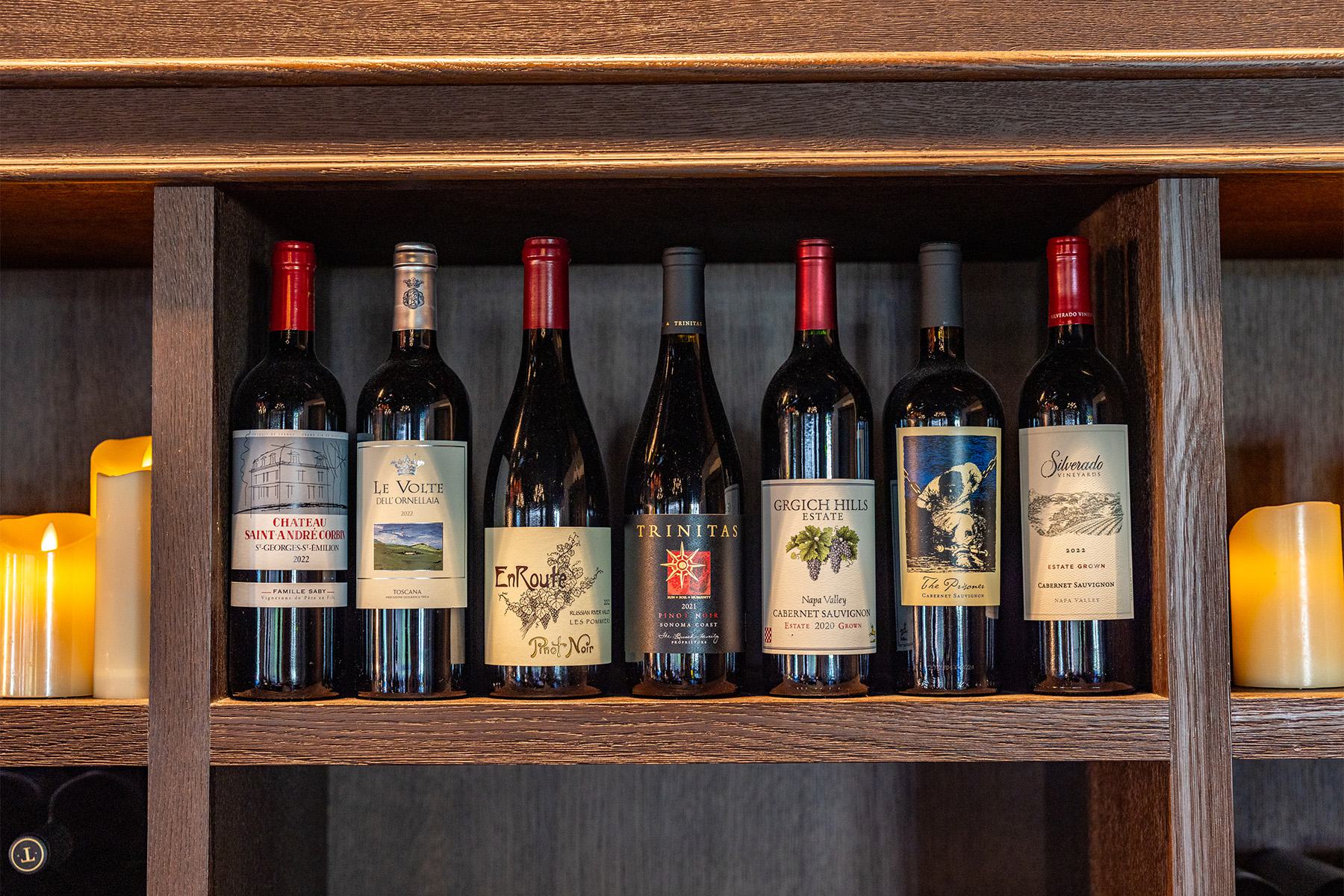 Seven bottles of red wine are displayed upright on a wooden shelf, with several unlit pillar candles positioned on either side—a perfect scene to celebrate restaurant month in Napa.